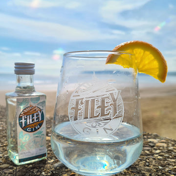 Filey branded gin tumbler glass on beach with miniature bottle of filey signature gin e